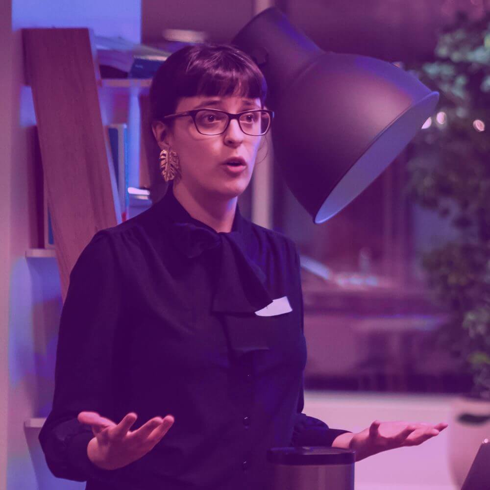 It's me, while presenting a talk at a womendotcode belgium meetup! I'm a white, feminine non-binary person with dark hair. I'm wearing a black blouse, large golden earrings in the shape of a leaf, and dark rimmed glasses.